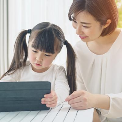 An image of a mother and daughter sitting at a table and looking at a smart tablet together. The daughter is on the left, holding the tablet. The mother is on the right supporting her exploration. The setting is bright, illuminated by natural light in the setting.